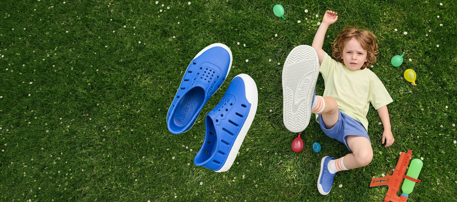 A child lies on green grass with colorful toy balls and a water gun nearby, while a pair of bright blue clog-style shoes with white soles floats beside them. The child is wearing matching blue shoes and striped socks.